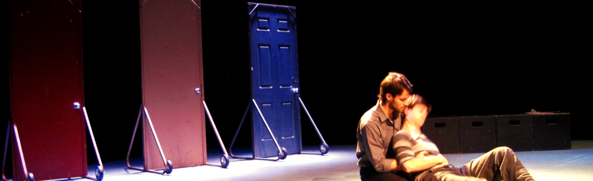 A man holds another man in his arms. They sit on the floor in front of three free-standing doors.